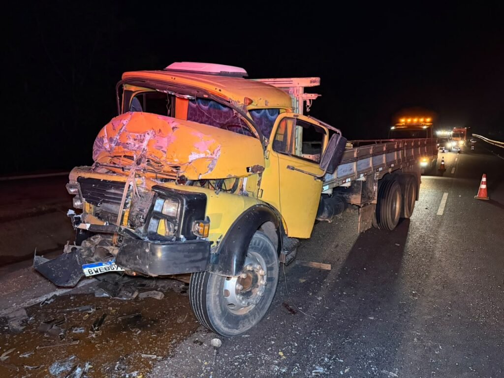 Caminhão amarelo com a frente e a cabine destruídas na beira da rodovia à noite
