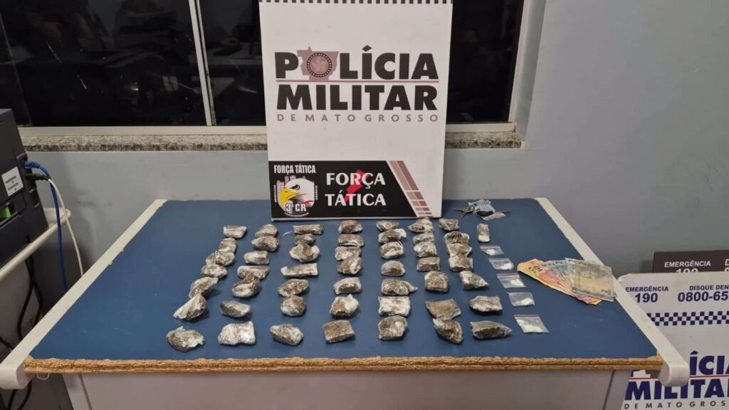 Porções de drogas e cédulas de dinheiro expostas sobre uma mesa, com identificação da Polícia Militar ao fundo