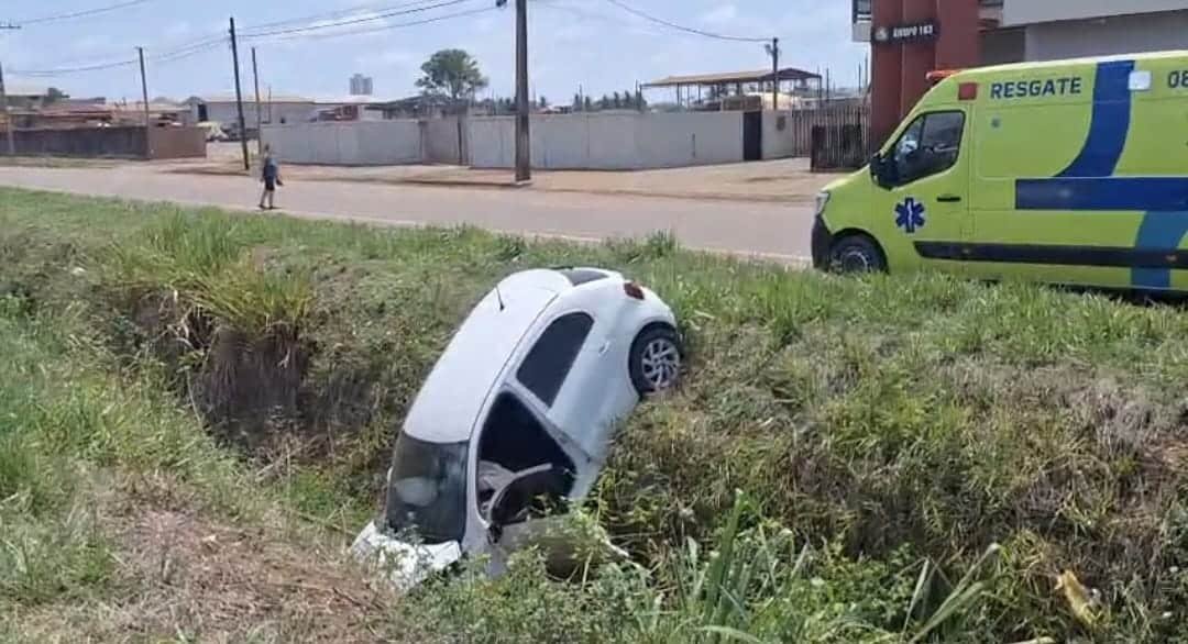 Carro branco dentro de vala gramada às margens de rodovia, com ambulância de resgate ao fundo